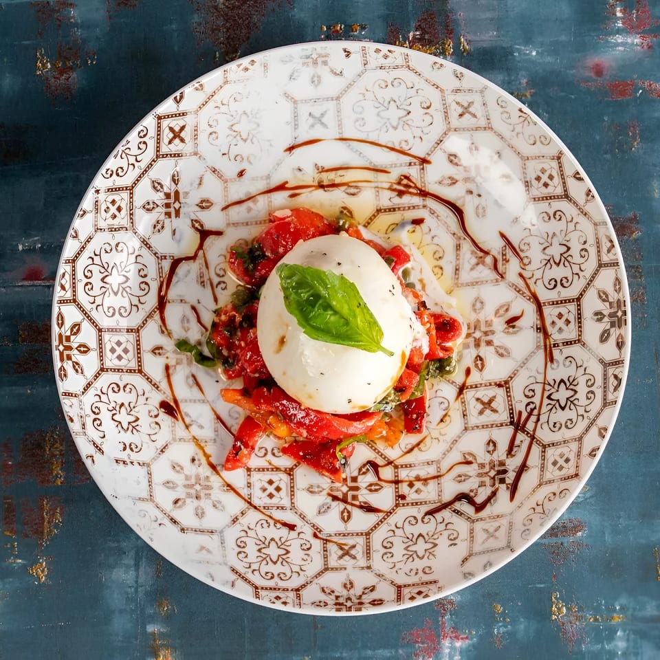 Burrata cheese with roasted red peppers, basil, and balsamic glaze on a decorative plate.