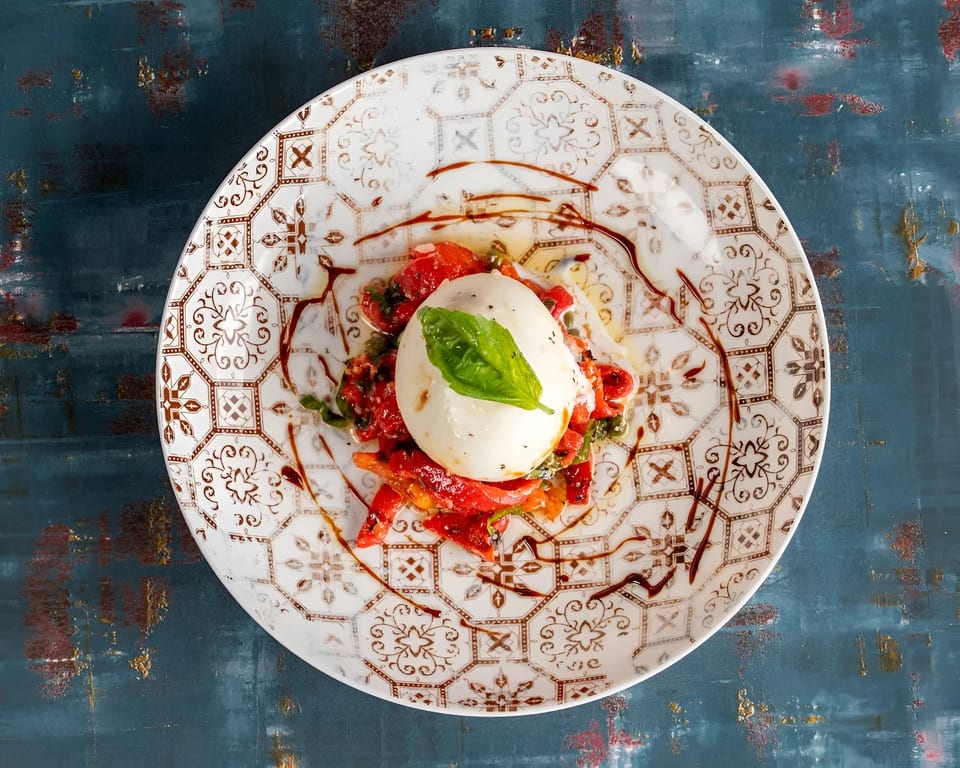 Burrata cheese with roasted red peppers, basil, and balsamic glaze on a decorative plate.
