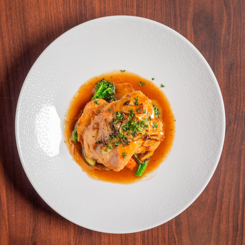 Plated chicken topped with mushrooms and herbs, served with vegetables in brown gravy on a white plate.