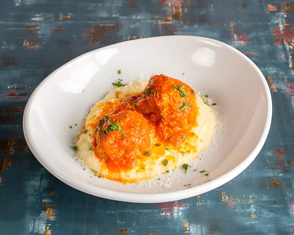 Two meatballs in red sauce on a bed of polenta, garnished with parsley and parmesan cheese.