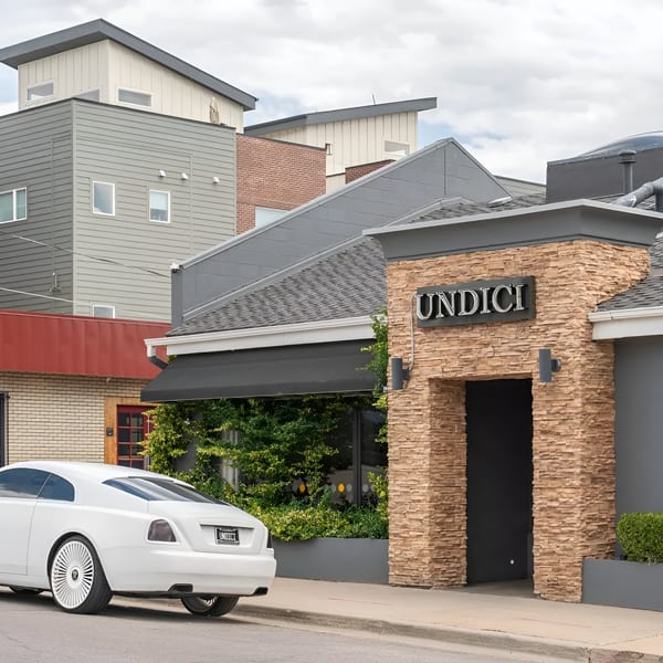 A white luxury car parked outside Undici restaurant, with modern buildings in the background.