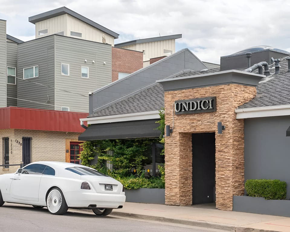 A white luxury car parked outside Undici restaurant, with modern buildings in the background.