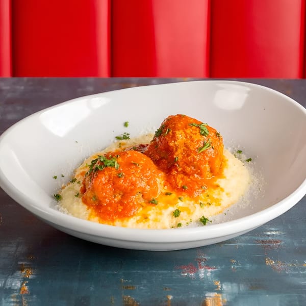 Two meatballs with red sauce and parsley on a bed of polenta in a white bowl.