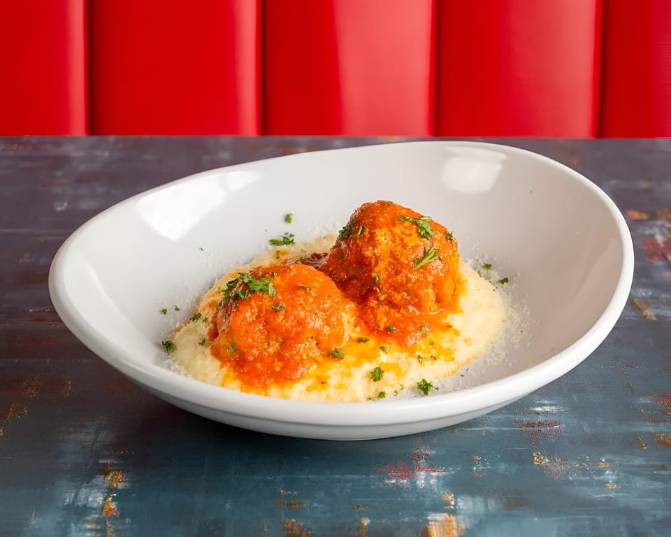 Two meatballs with red sauce and parsley on a bed of polenta in a white bowl.