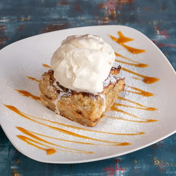 Bread pudding topped with vanilla ice cream, powdered sugar, and caramel drizzle on a white plate.