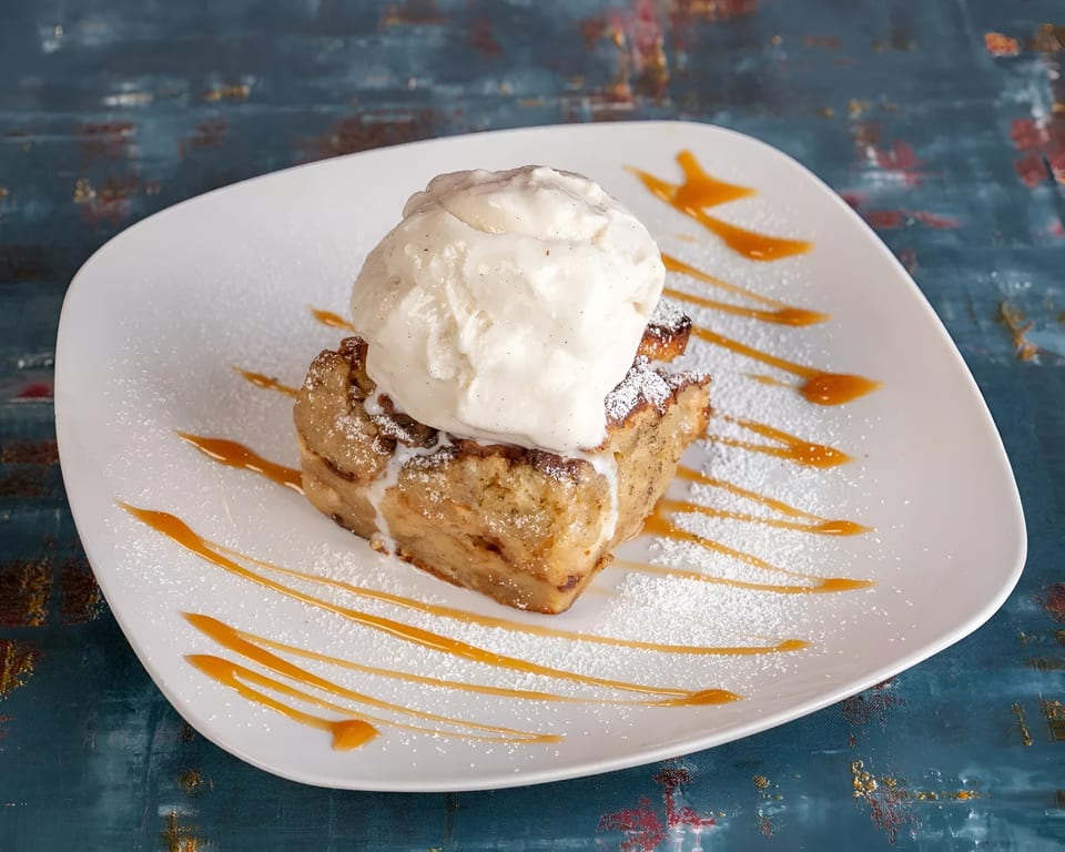 Bread pudding topped with vanilla ice cream, powdered sugar, and caramel drizzle on a white plate.