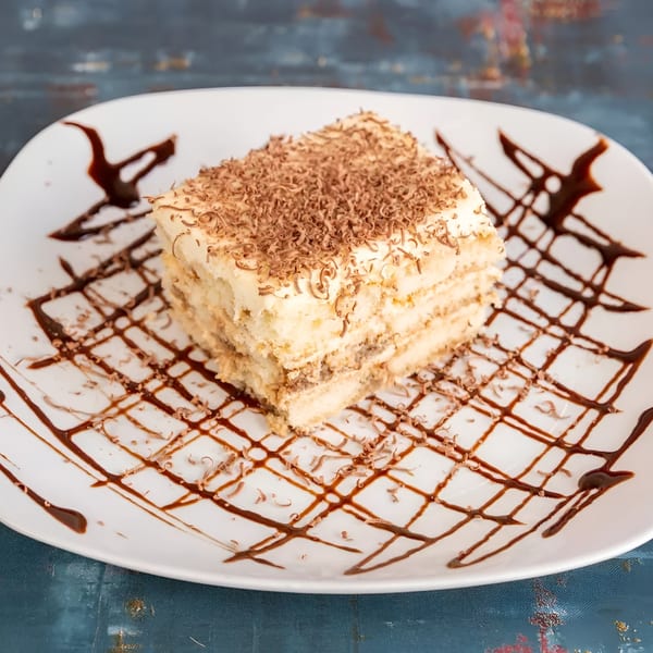 A slice of tiramisu with chocolate shavings and chocolate syrup on a white plate.