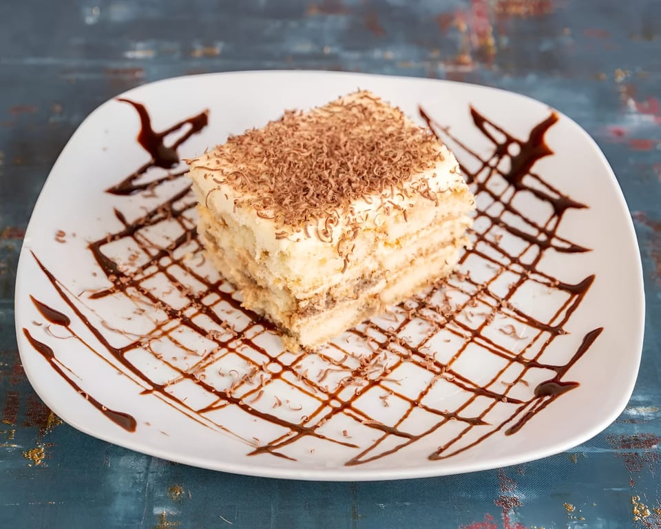 A slice of tiramisu with chocolate shavings and chocolate syrup on a white plate.