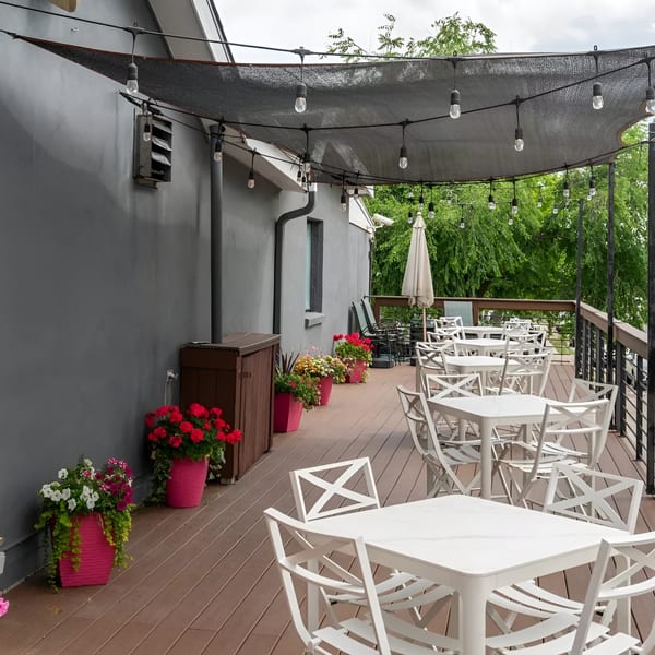 Outdoor restaurant patio with white tables and chairs, string lights, and potted flowers under a dark canopy.