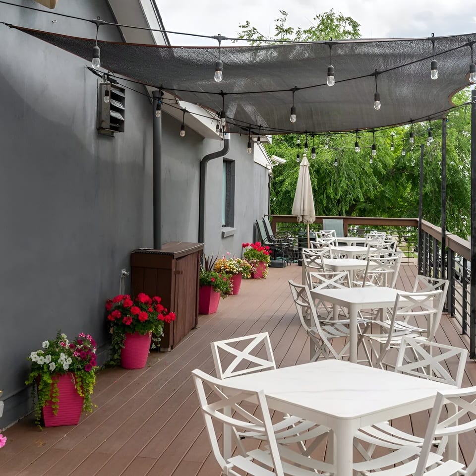 Outdoor restaurant patio with white tables and chairs, string lights, and potted flowers under a dark canopy.
