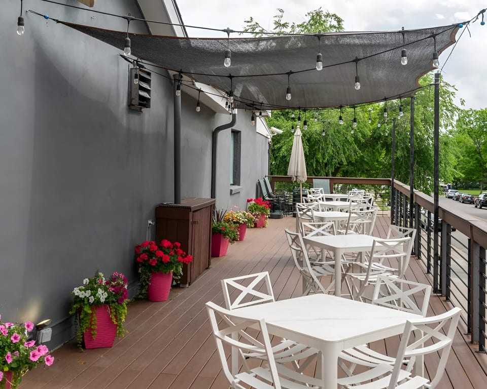 Outdoor restaurant patio with white tables and chairs, string lights, and potted flowers under a dark canopy.
