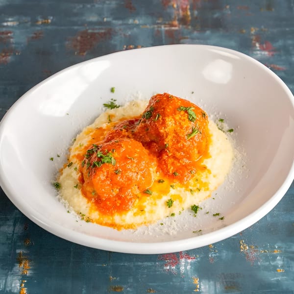 Two meatballs in red sauce over polenta, garnished with parsley and parmesan, in a white bowl.